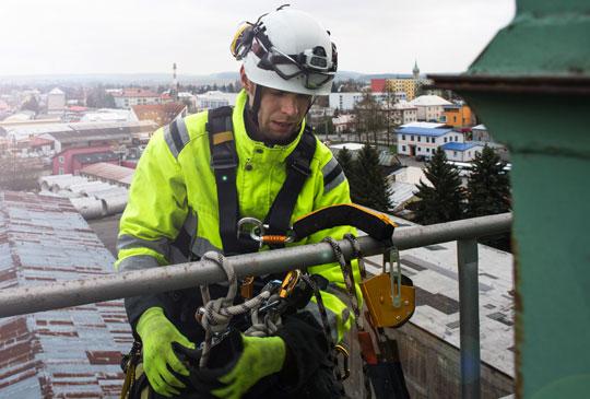 En person i varselkläder och skyddshjälm utför arbete på hög höjd med säkerhetssele och rep fastsatta i ett räcke.