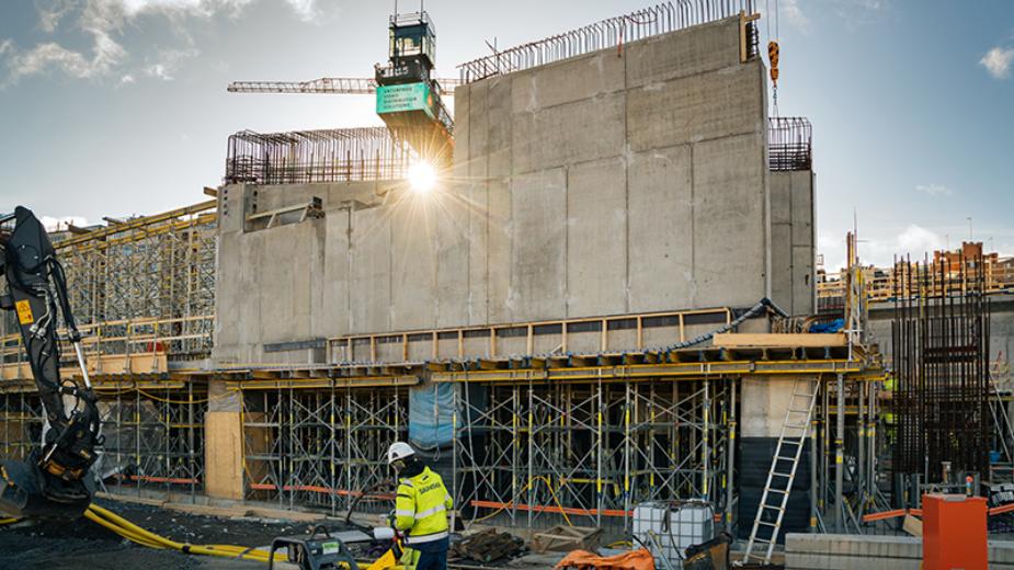 En byggarbetsplats med en stor betongvägg, byggnadsställningar och en byggarbetare i gula varselkläder i förgrunden.