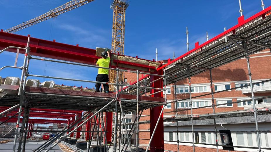 En byggarbetare i gul skyddsväst monterar isolering på en röd stålbalk på en byggställning.