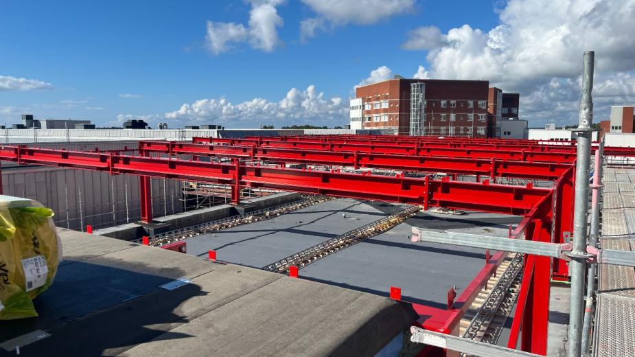Röda stålbalkar monterade över en byggnad under ett pågående arbete utomhus.