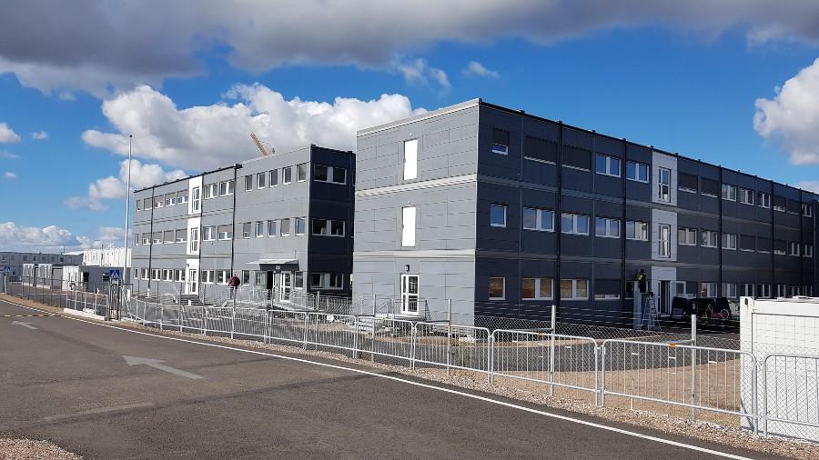 Gråa modulbyggnader i tre våningar längs en asfalterad väg under en blå himmel med vita moln. Ett metallstängsel finns framför byggnaderna.