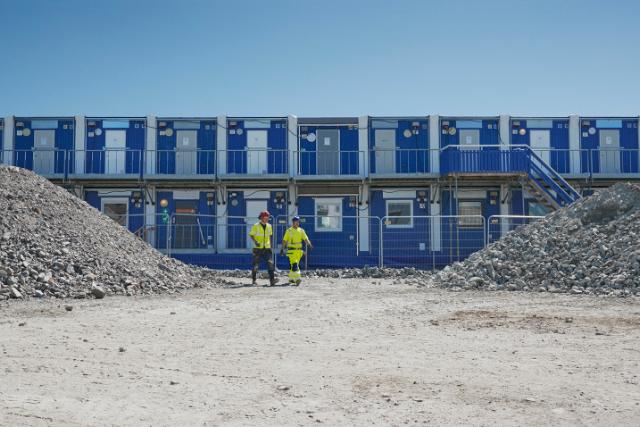 Två byggarbetare i gula varselkläder står på en grusig byggarbetsplats framför en blå bodetablering, med stora grushögar på båda sidor.